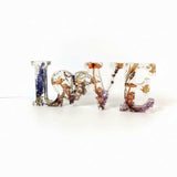 Resin mushroom decor spells LOVE in clear resin letters filled with dried mushrooms, ferns, and gold flecks, with a heart-shaped O.