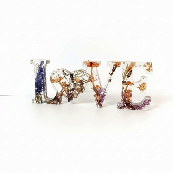 Resin mushroom decor spells LOVE in clear resin letters filled with dried mushrooms, ferns, and gold flecks, with a heart-shaped O.