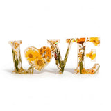 Pressed flower resin love sign in clear resin, spelling LOVE with yellow wildflowers and a floral heart-shaped O