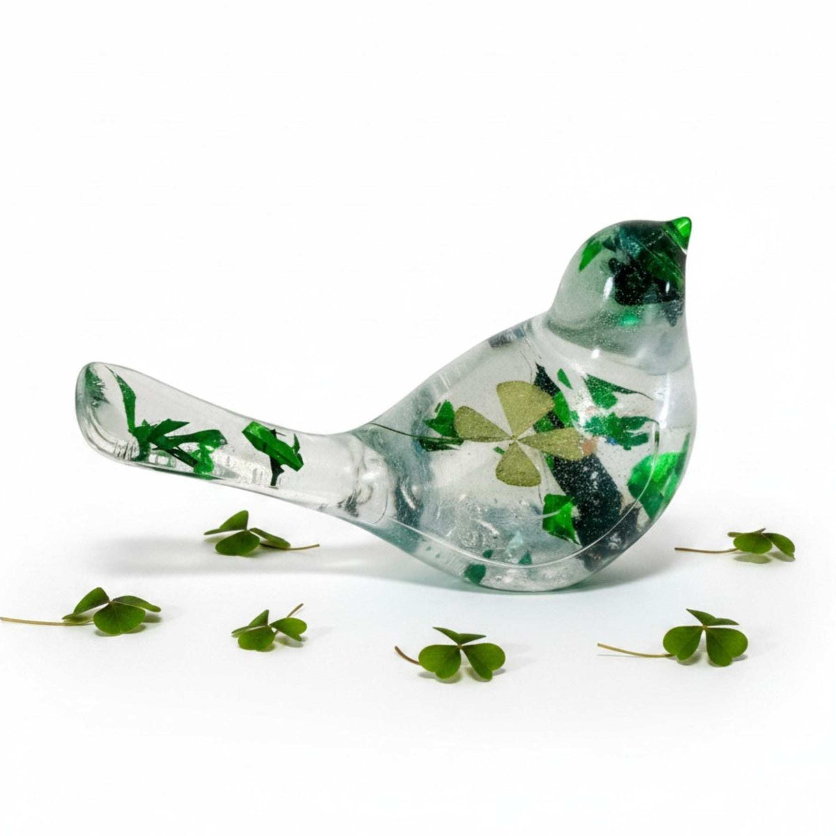 Clear resin bird figurine filled with real green four leaf clovers, vibrant green botanicals, and gold flakes, surrounded by fresh clover leaves on a white background.