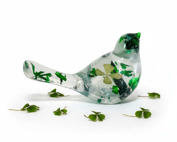 Clear resin bird figurine filled with real green four leaf clovers, vibrant green botanicals, and gold flakes, surrounded by fresh clover leaves on a white background.