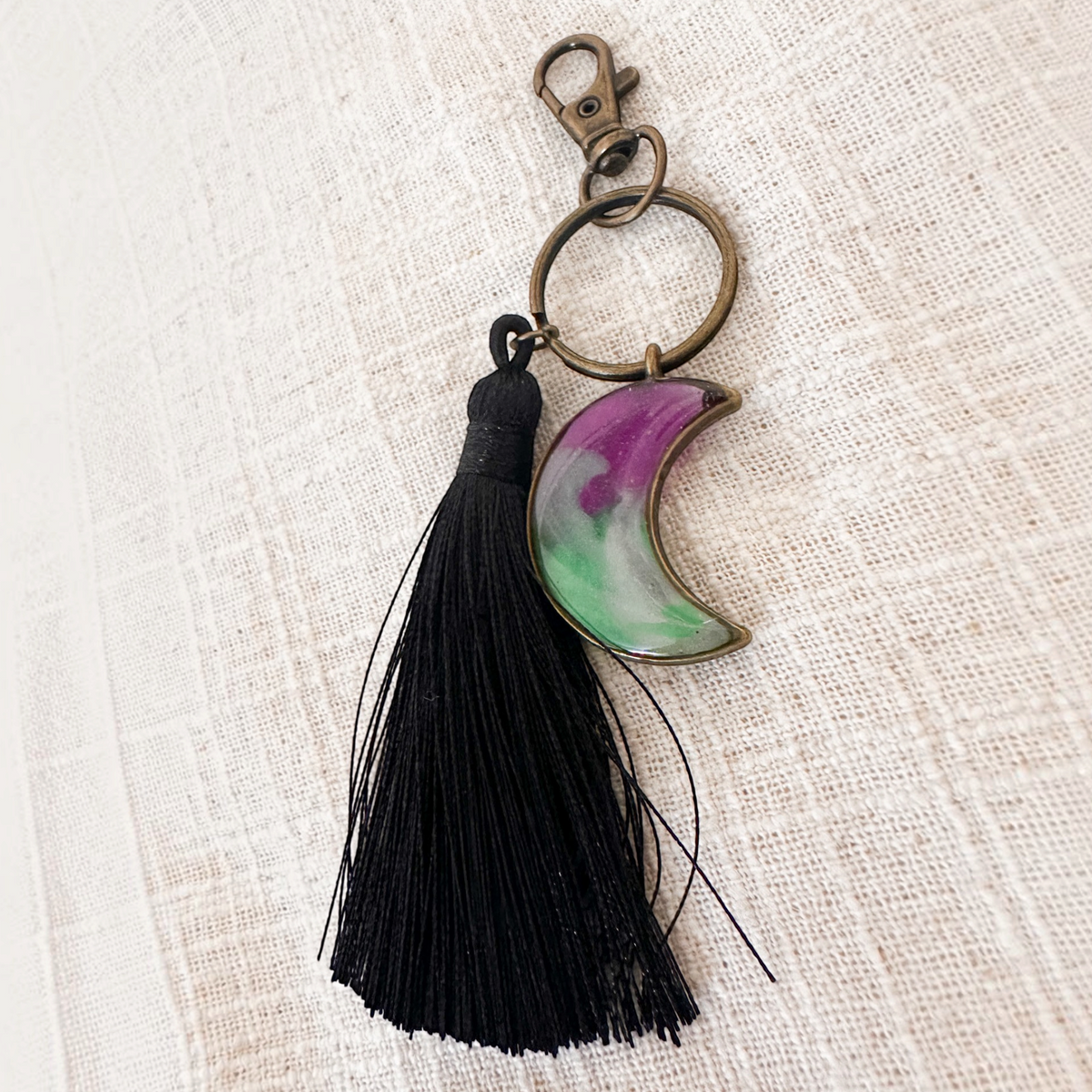 Luna Glow crescent moon keychain in translucent purple and green swirled resin, with an antique bronze clasp and black tassel
