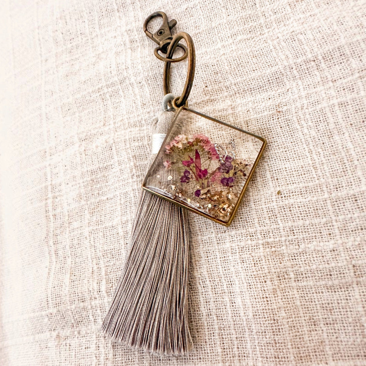 Meadow Butterfly pressed flower bag charm in clear resin, with pink and purple blooms, bronze frame and grey tassel