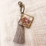 Meadow Butterfly pressed flower bag charm in clear resin, with pink and purple blooms, bronze frame and grey tassel