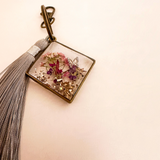 Meadow Butterfly resin bag charm with pressed pink and purple flowers, gold flakes, bronze frame and grey tassel