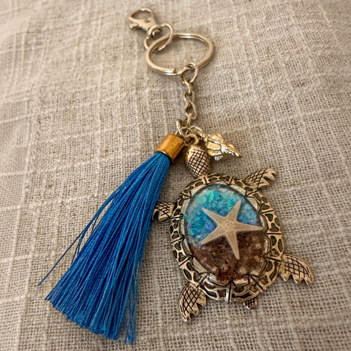 Ocean Keeper sea turtle keychain with antique bronze tone metal, blue tassel, and clear resin holding a starfish and sand