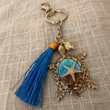 Ocean Keeper sea turtle keychain with antique bronze tone metal, blue tassel, and clear resin holding a starfish and sand