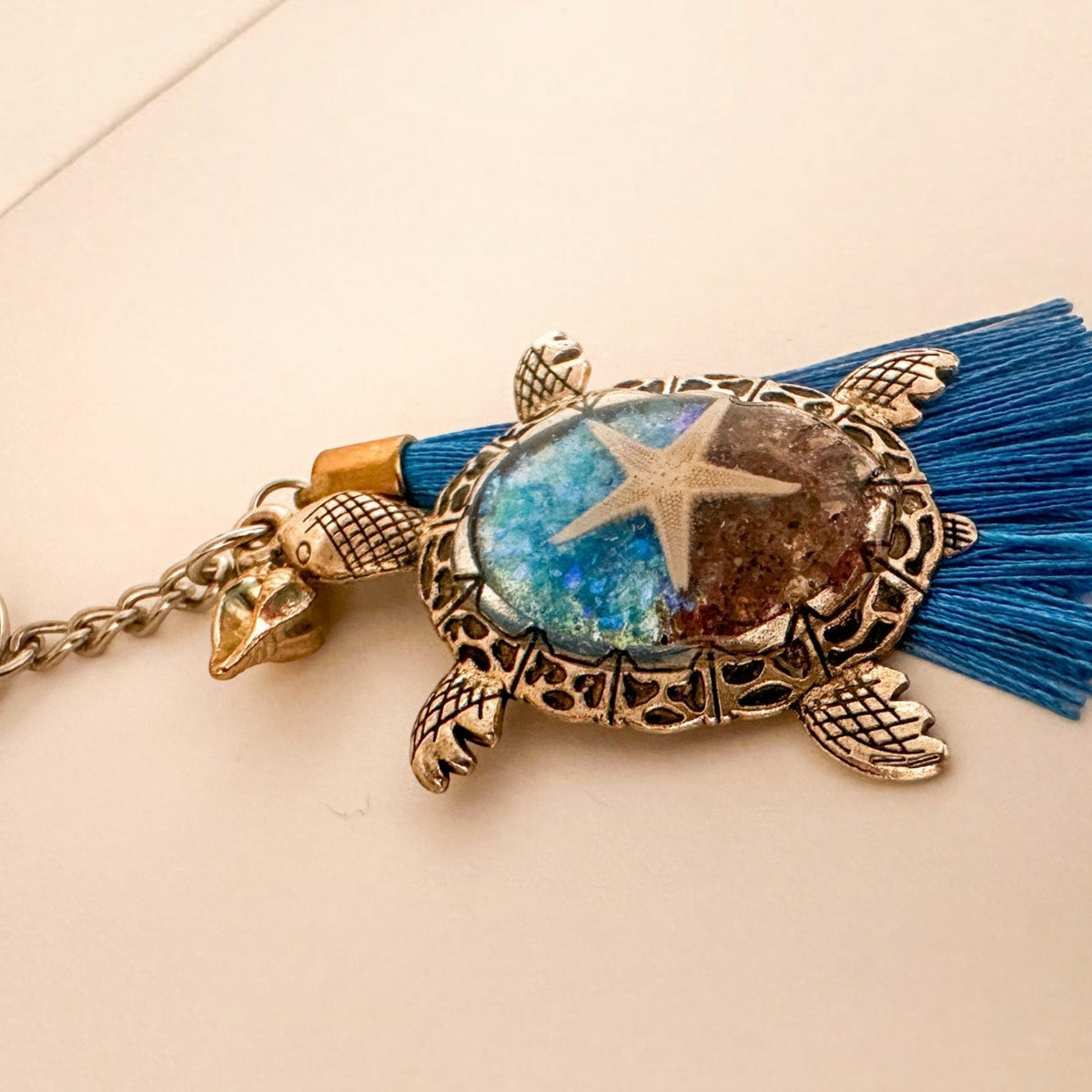 Ocean Keeper turtle keychain with an antique bronze metal frame, blue tassel, and glossy beach scene featuring a starfish