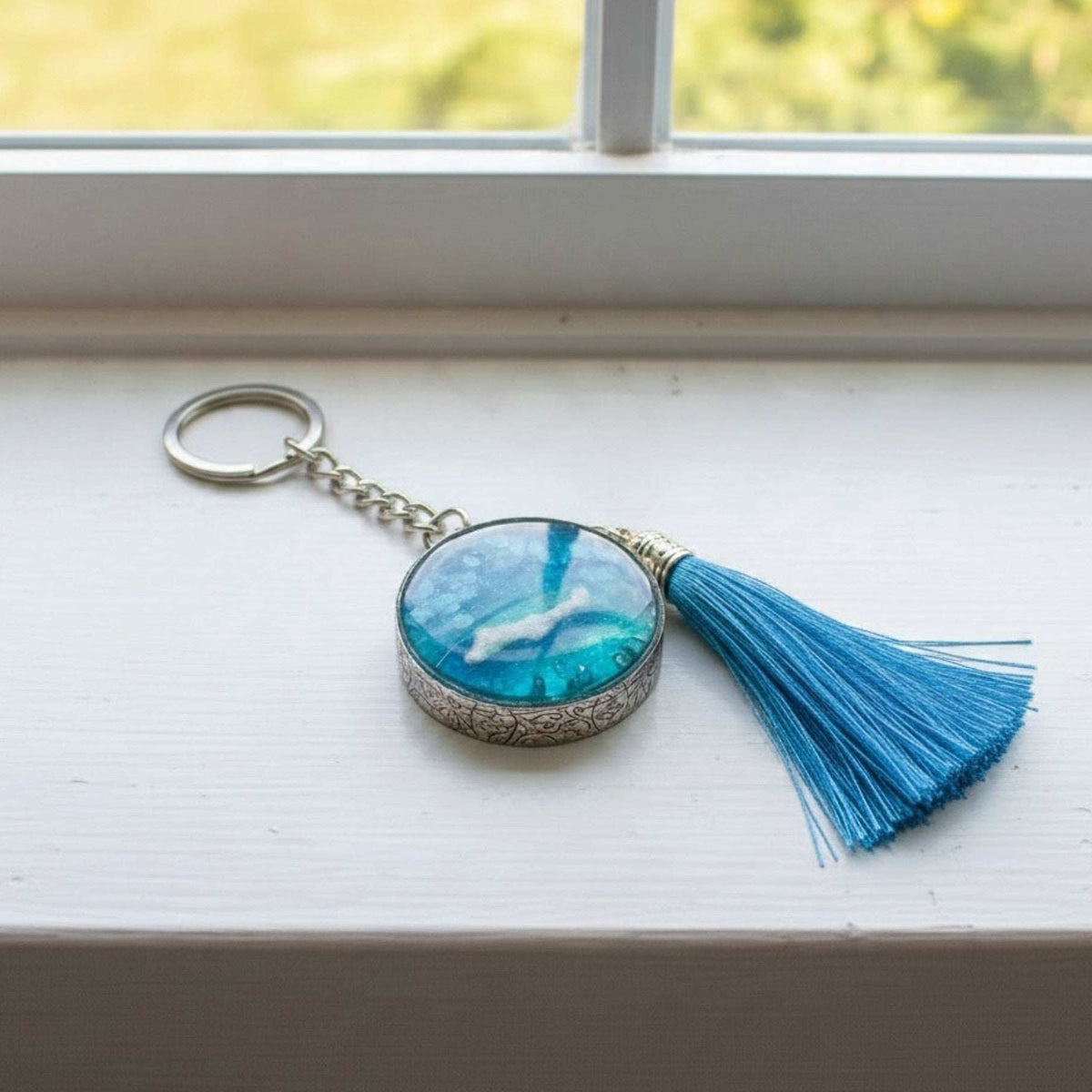 Glow in the dark ocean keychain with layered turquoise resin, white wave detail, ornate silver frame and blue tassel.