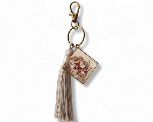 Pressed flower resin bag charm with clear square resin, pink and purple pressed blooms, bronze hardware and a grey tassel.