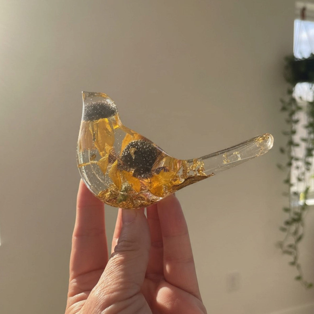 black eyed susan resin bird in clear resin with amber-yellow dried flowers, dark centres, and gold leaf accents along its tail and base
