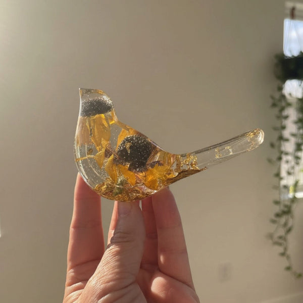 black eyed susan resin bird in clear resin with amber-yellow dried flowers, dark centres, and gold leaf accents along its tail and base
