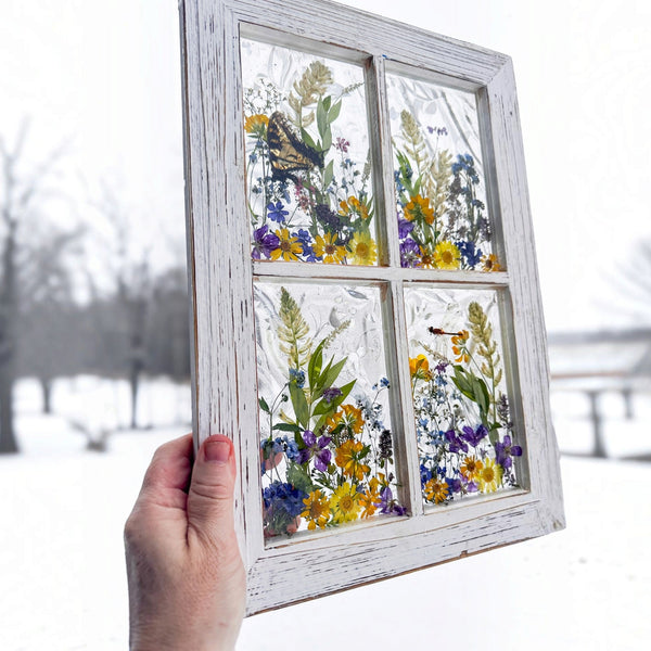 Pressed flower window art hanging on a wall in its white distressed wood frame