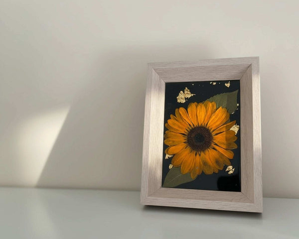 Pressed sunflower frame art with a dimensional orange bloom and green leaves on black with gold flecks, in a light wood frame