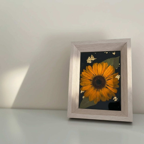Pressed sunflower frame art with a dimensional orange bloom and green leaves on black with gold flecks, in a light wood frame