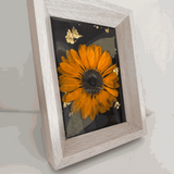 Pressed sunflower frame art with a dimensional orange bloom, muted green leaves and gold flecks on black resin in a pale wood frame
