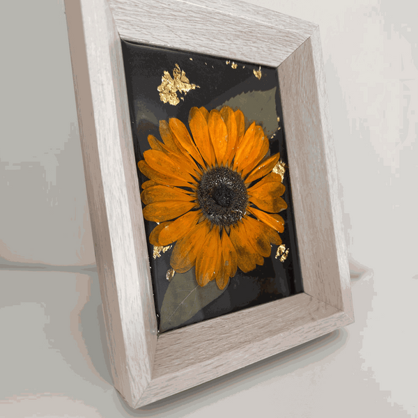 Pressed sunflower frame art with a dimensional orange bloom, muted green leaves and gold flecks on black resin in a pale wood frame