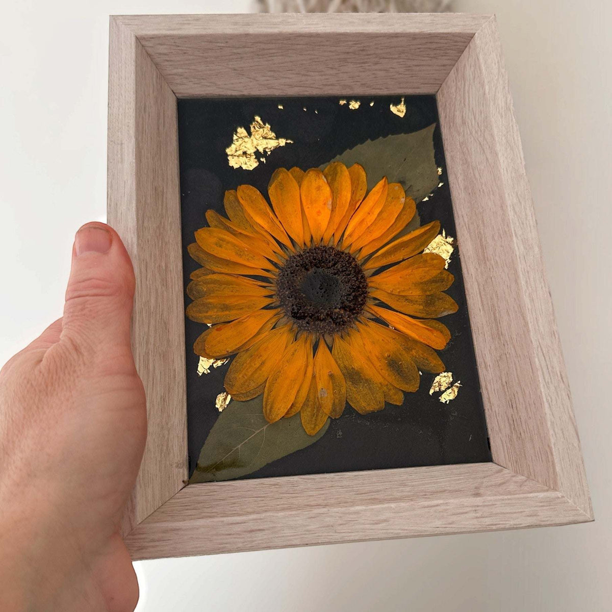Pressed sunflower frame art with a three-dimensional orange bloom and green leaves on black with gold flecks, in a pale wood frame.