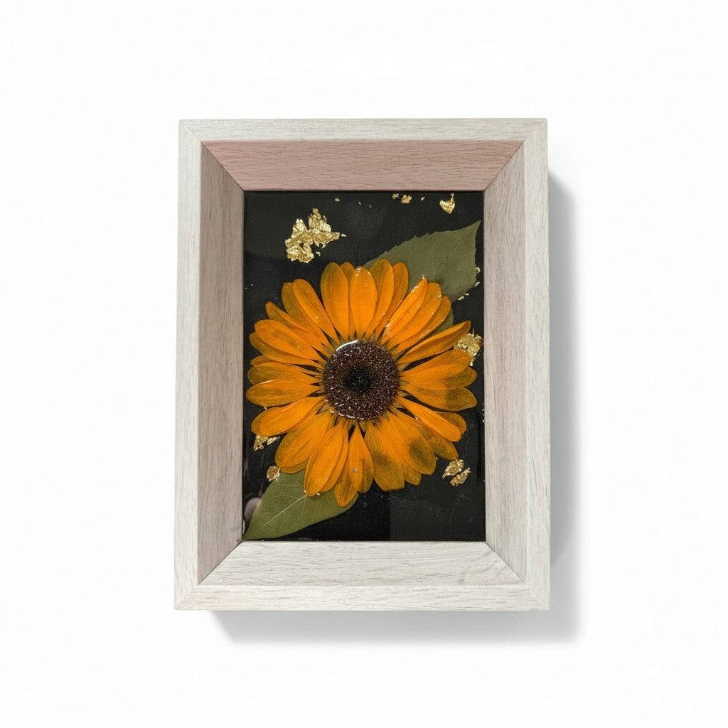 Pressed sunflower frame art with a dimensional orange bloom, green leaves, gold flecks on black, and a pale wood frame