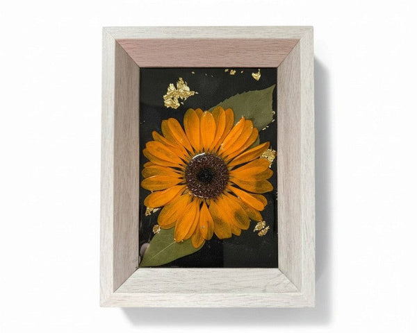 Pressed sunflower frame art with a dimensional orange bloom, green leaves, gold flecks on black, and a pale wood frame