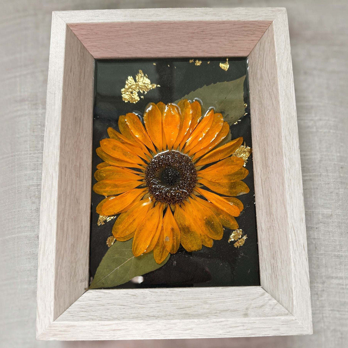 Pressed sunflower frame art with a dimensional orange bloom and green leaves on black with gold flecks, in a pale wood frame.