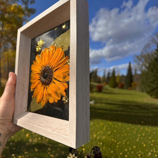 Pressed sunflower frame art with a three-dimensional orange bloom in a clear resin panel, black and gold details, and a pale wood frame.
