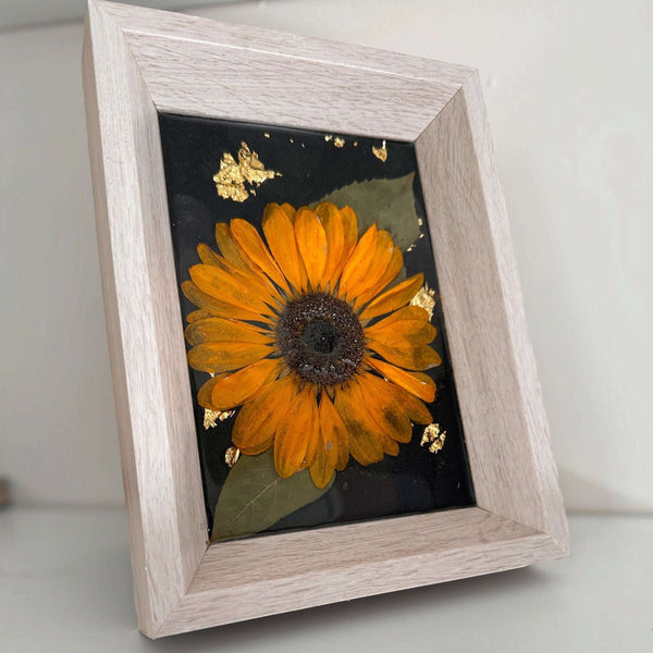 pressed sunflower frame art with a vivid orange three-dimensional bloom, green leaf and gold flecks on black in a pale wood frame