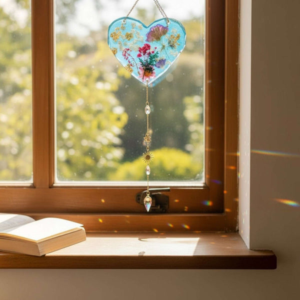Glow in the dark heart suncatcher made of blue resin with pressed pink and purple wildflowers, gold leaf, and hanging crystal accents