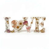 Whispering Meadow LOVE resin word art with white daisies, blush pink and purple wildflowers, bunny tail, and gold leaf flakes